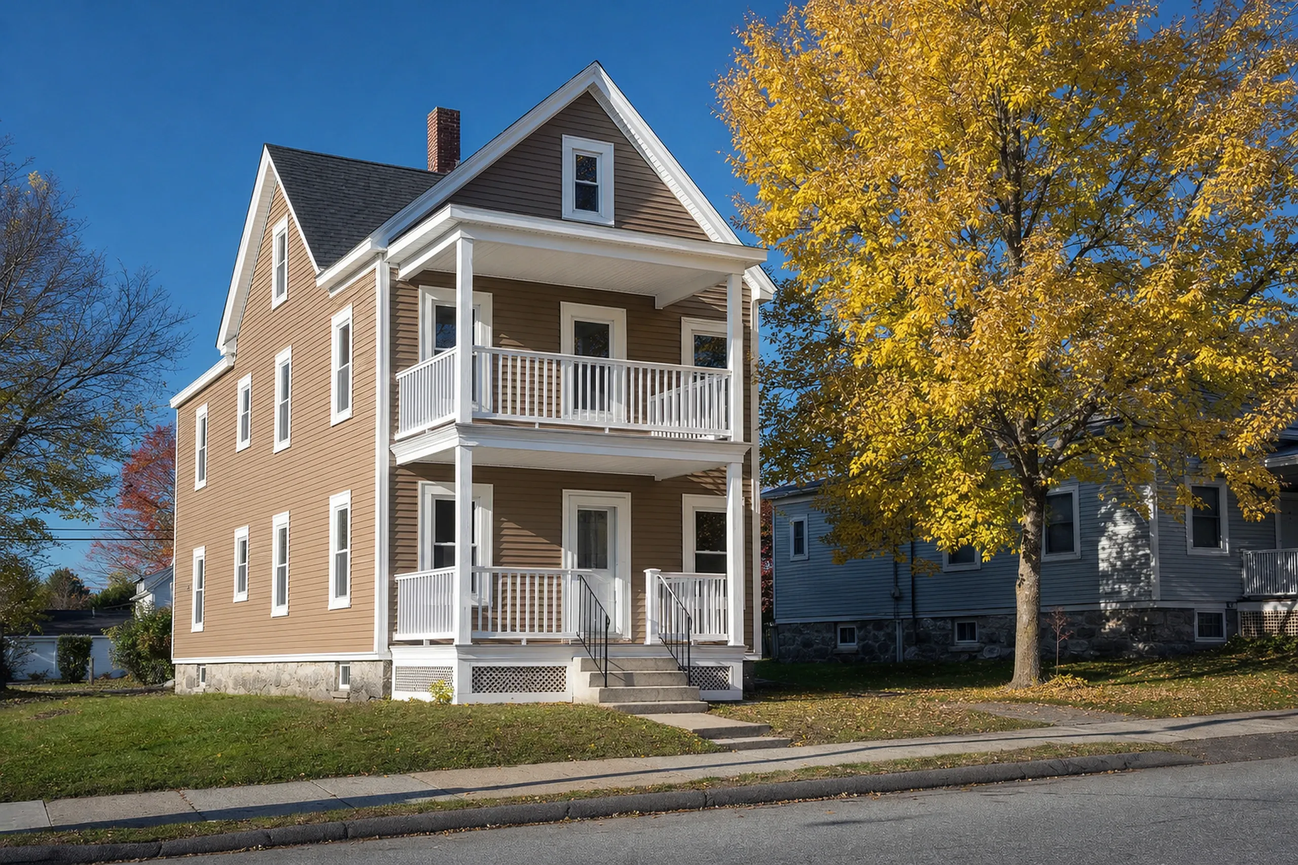 Finished re-side in Lewiston, ME: a two-and-a-half-story wood-frame two-family house with stacked front porches, new clay-colored vinyl lap siding, white corner boards and wide white window trim, a small city lawn and a maple in early fall