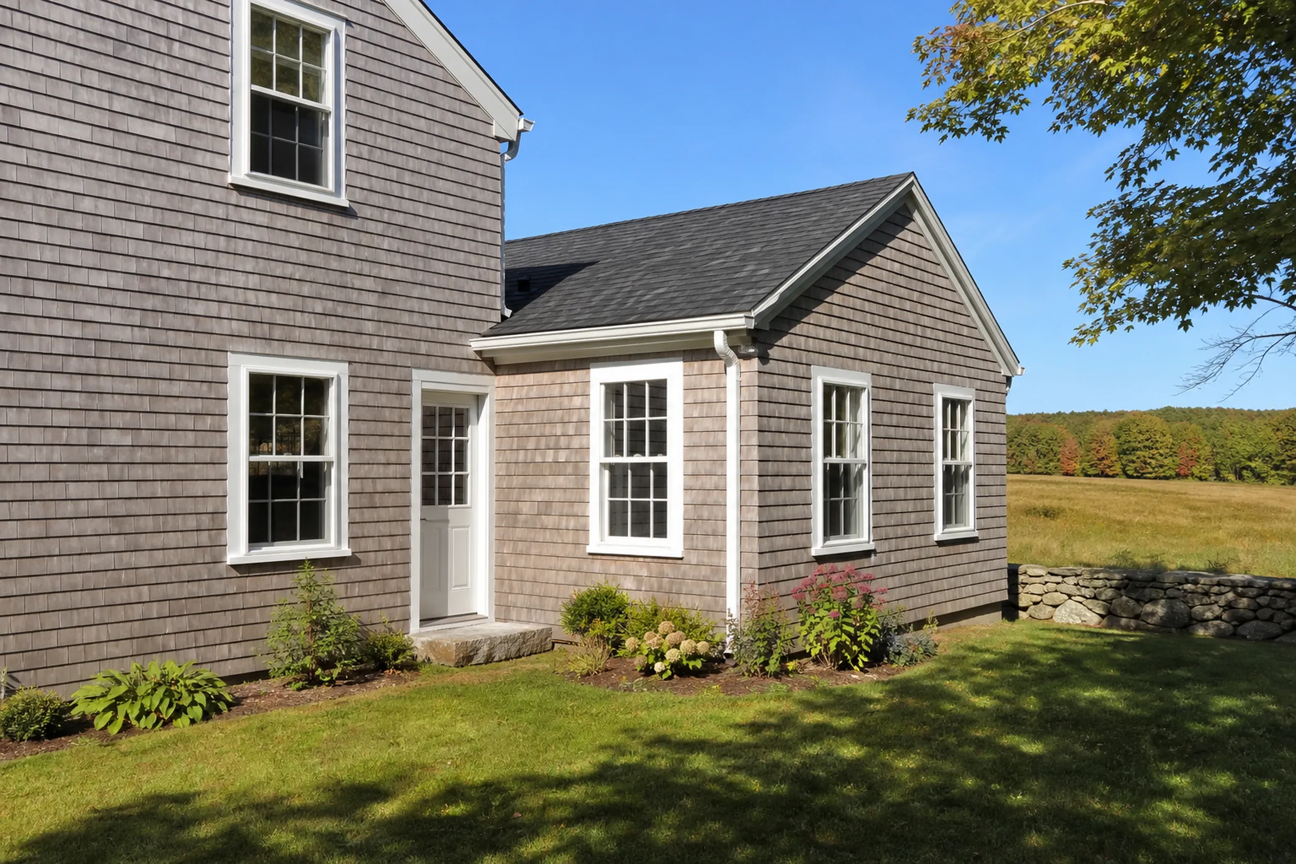 Gray-finished cedar shingles on a Lewiston-Auburn area farmhouse: cedar shingles in a soft gray semitransparent finish across the main house and its long single-story rear section, white trim, an open hayfield and maples behind, early fall.