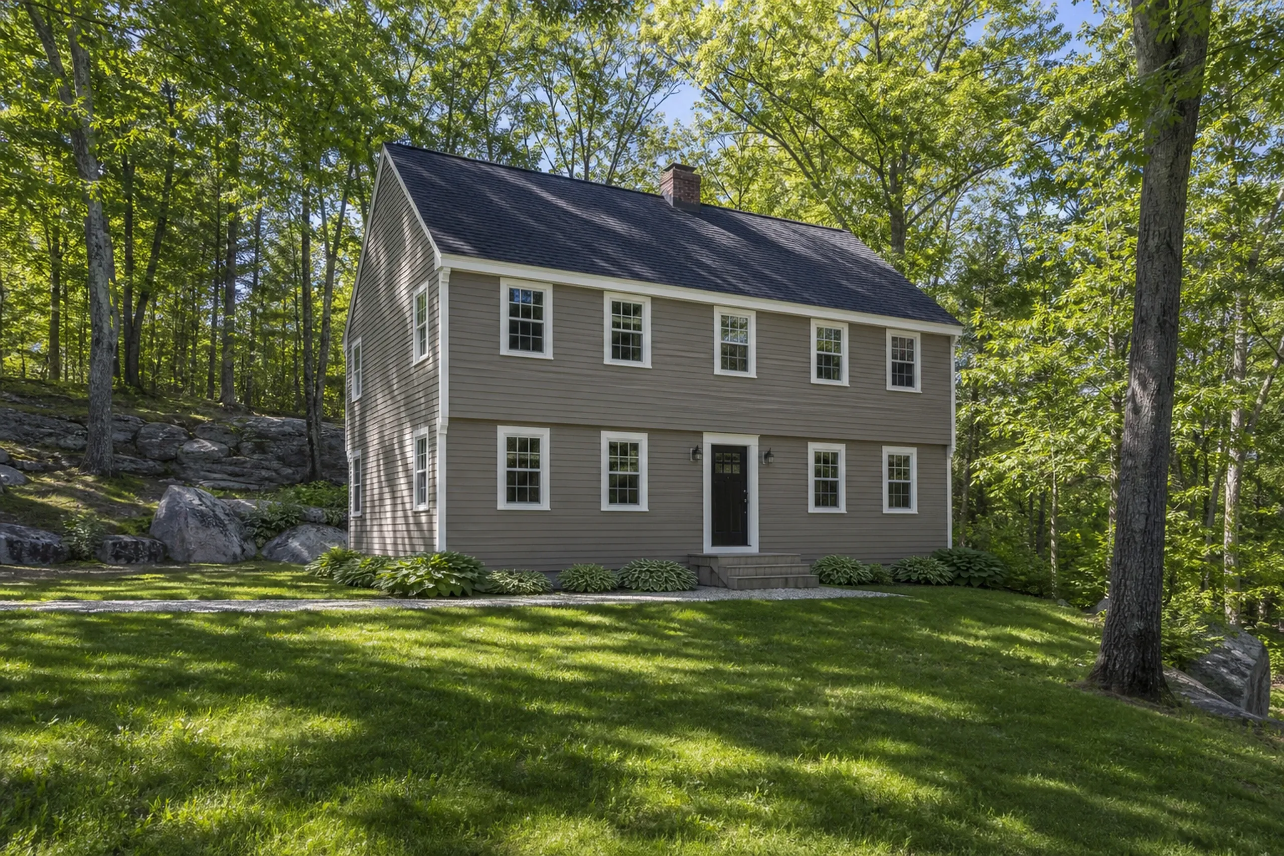 Re-sided Hardie garrison in the Lewiston-Auburn area: A re-sided garrison colonial on a wooded hillside lot, factory-finished fiber cement lap in warm taupe with white corner boards, white window casings and a black door, hostas along the foundation and sunlight through new oak leaves, bright early-summer afternoon light.