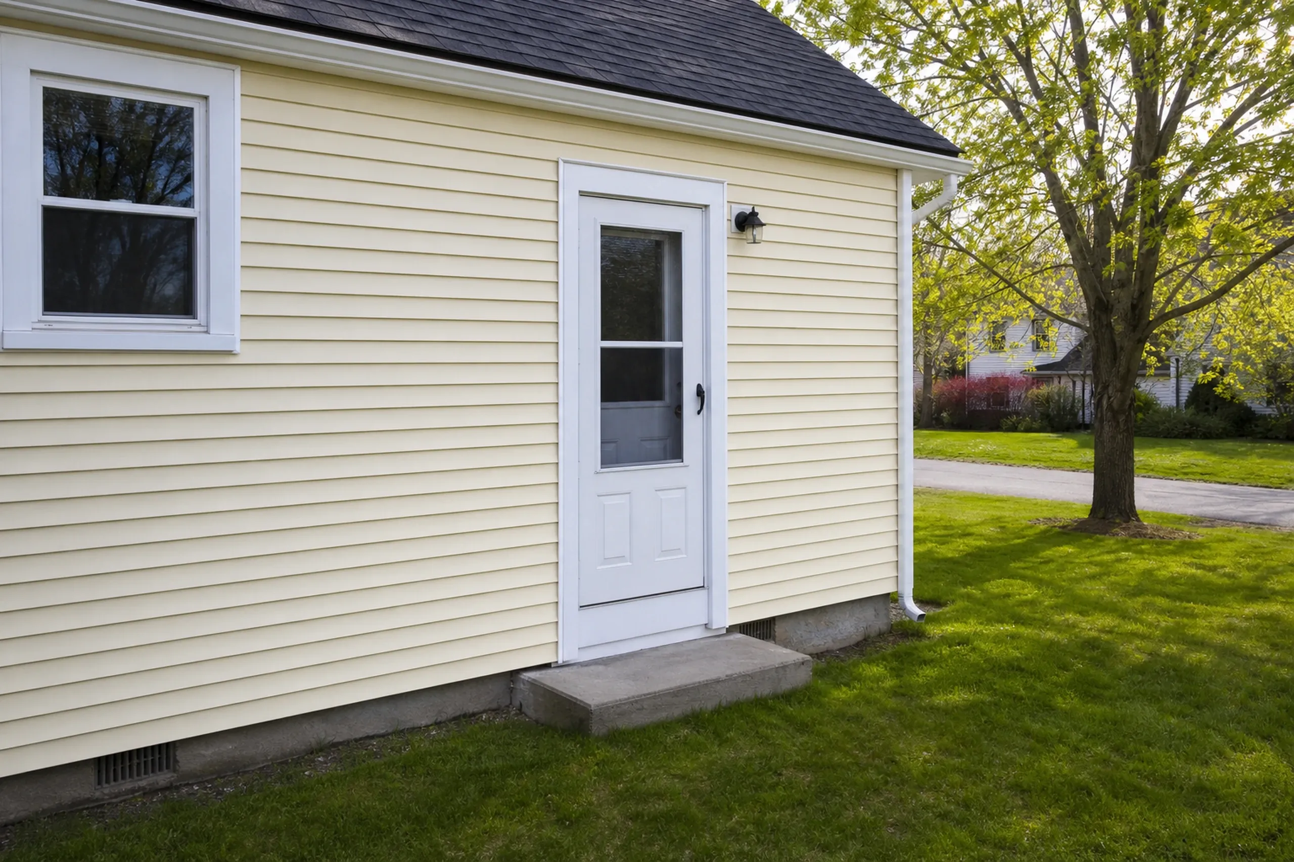 Siding repair on a 1950s Auburn house: a section of new pale yellow vinyl panels blended into the existing wall beside a side door, same profile and a close color match, a small lawn and a maple tree, mid-spring.