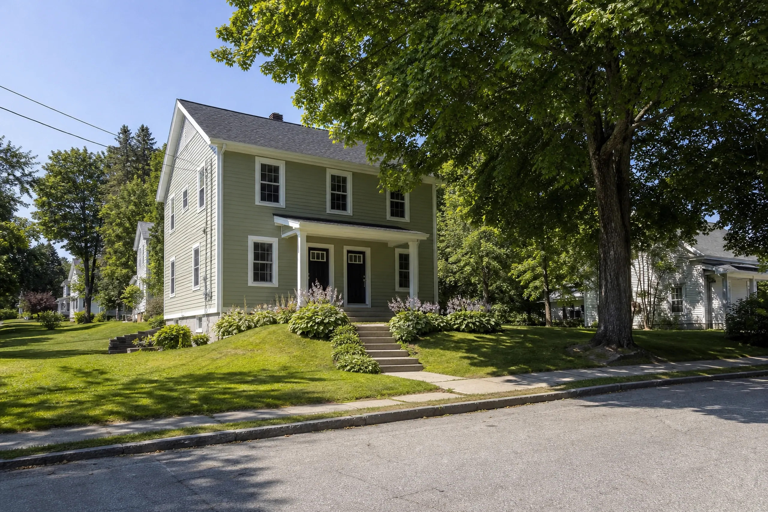 Re-sided Auburn mill-era duplex in early summer: A re-sided mill-era duplex on a tree-lined Auburn hill street, finished in muted sage fiber cement lap with white corner boards and trim, two matching front doors under a shared porch, flowering hostas by the steps and a sugar maple in full leaf, bright early-summer afternoon light.