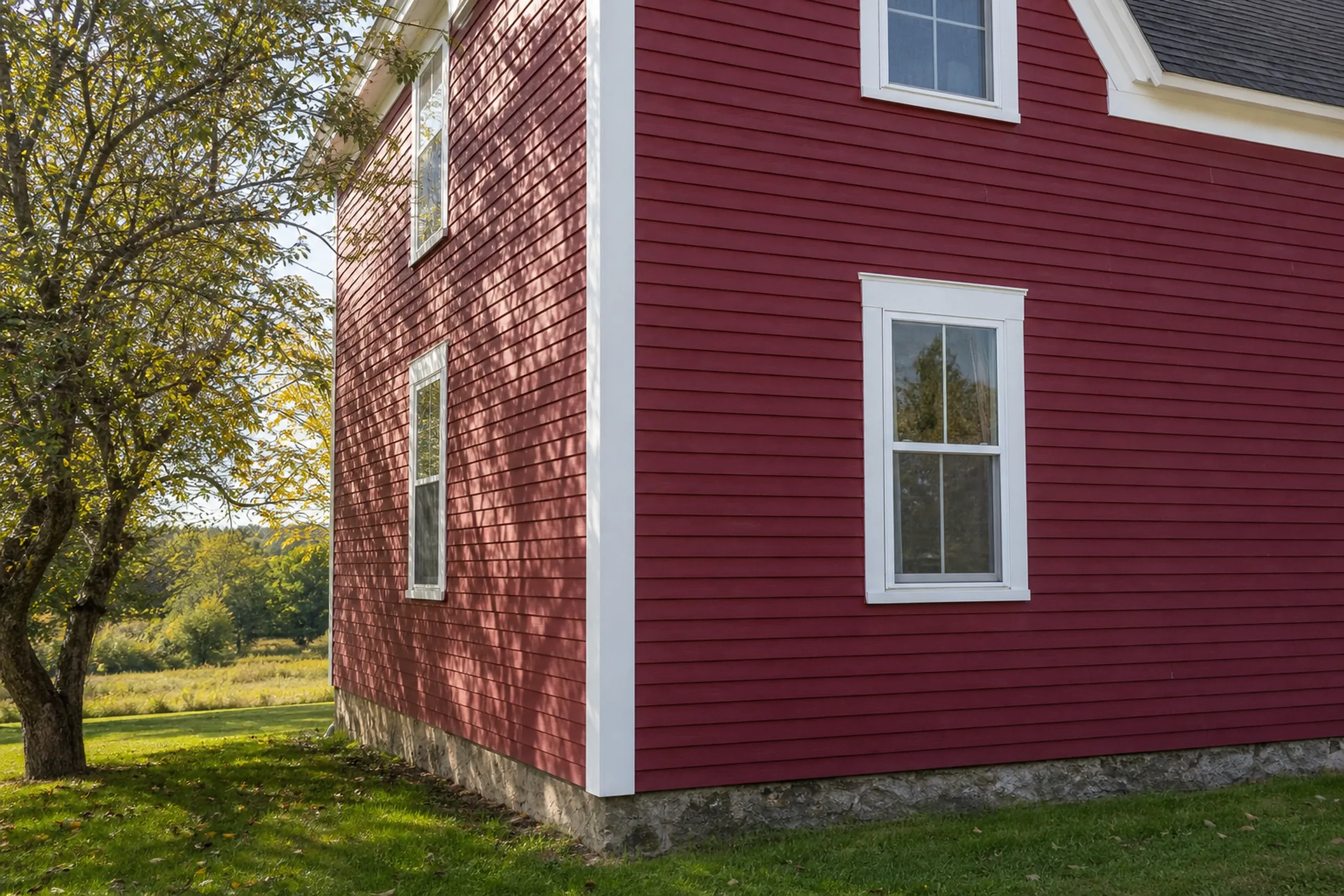 LP SmartSide on a Bowdoin farmhouse: close view of one corner of an old two-story farmhouse re-sided in barn-red painted engineered wood lap boards with a cedar-grain texture, a wide white corner board and window casing, an apple tree at the edge of the frame, early fall.