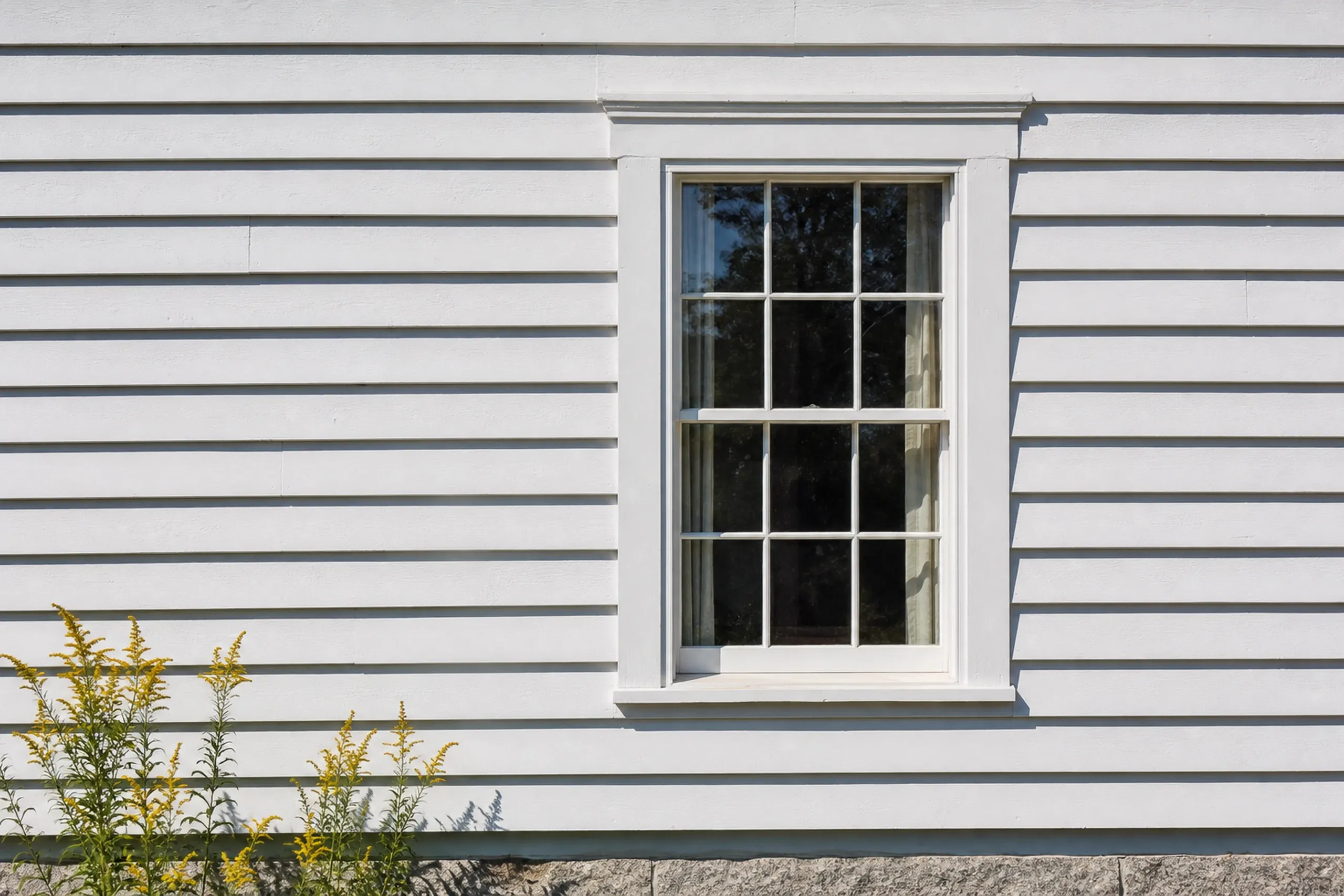 Clapboard repair in Buckfield Village: a weathered white clapboard wall with a few new primed boards pieced in at staggered end joints beside a window casing, late summer.