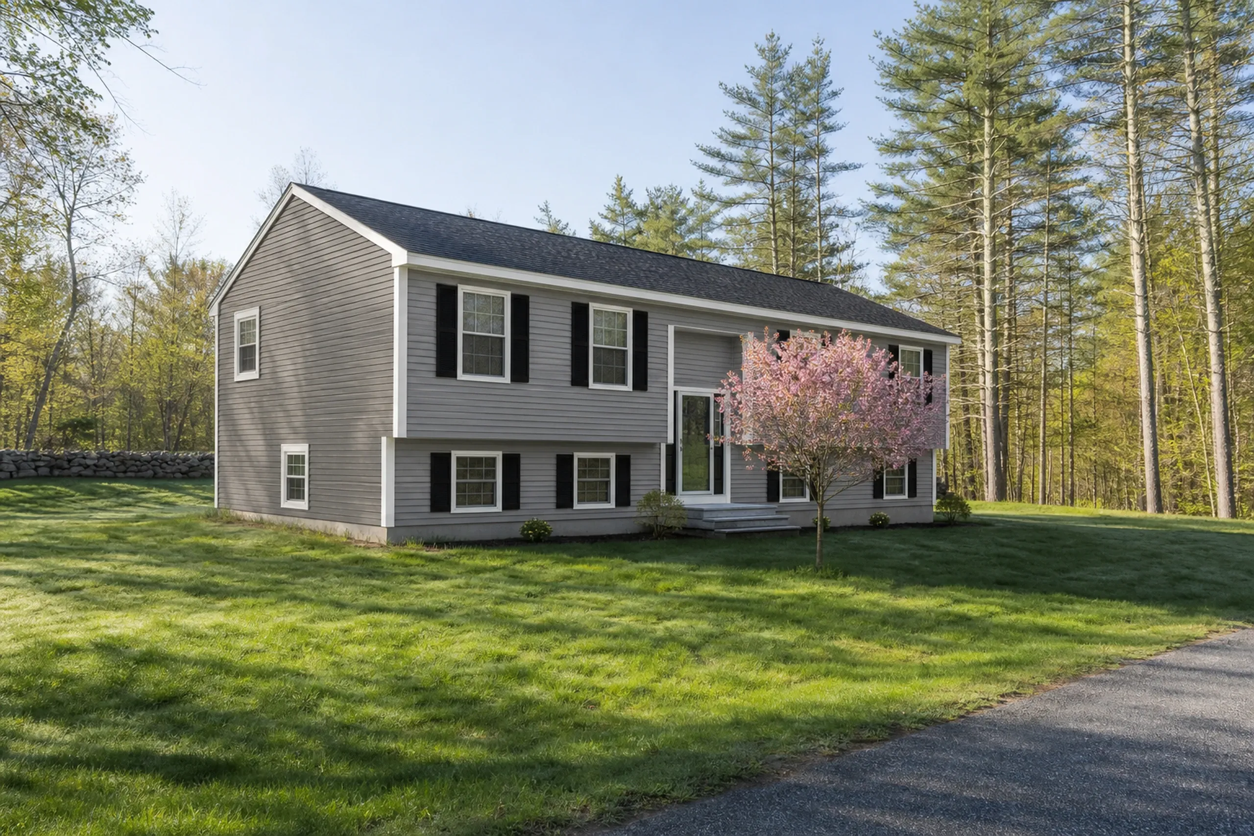 Re-sided Buckfield split-level in late spring: A re-sided 1980s split-level on a back road, mushroom-gray vinyl lap siding with white corner posts, white trim and black shutters, a flowering crabapple in pink bloom, a mown lawn running up to white pines and a stone wall, clear late-spring morning light.