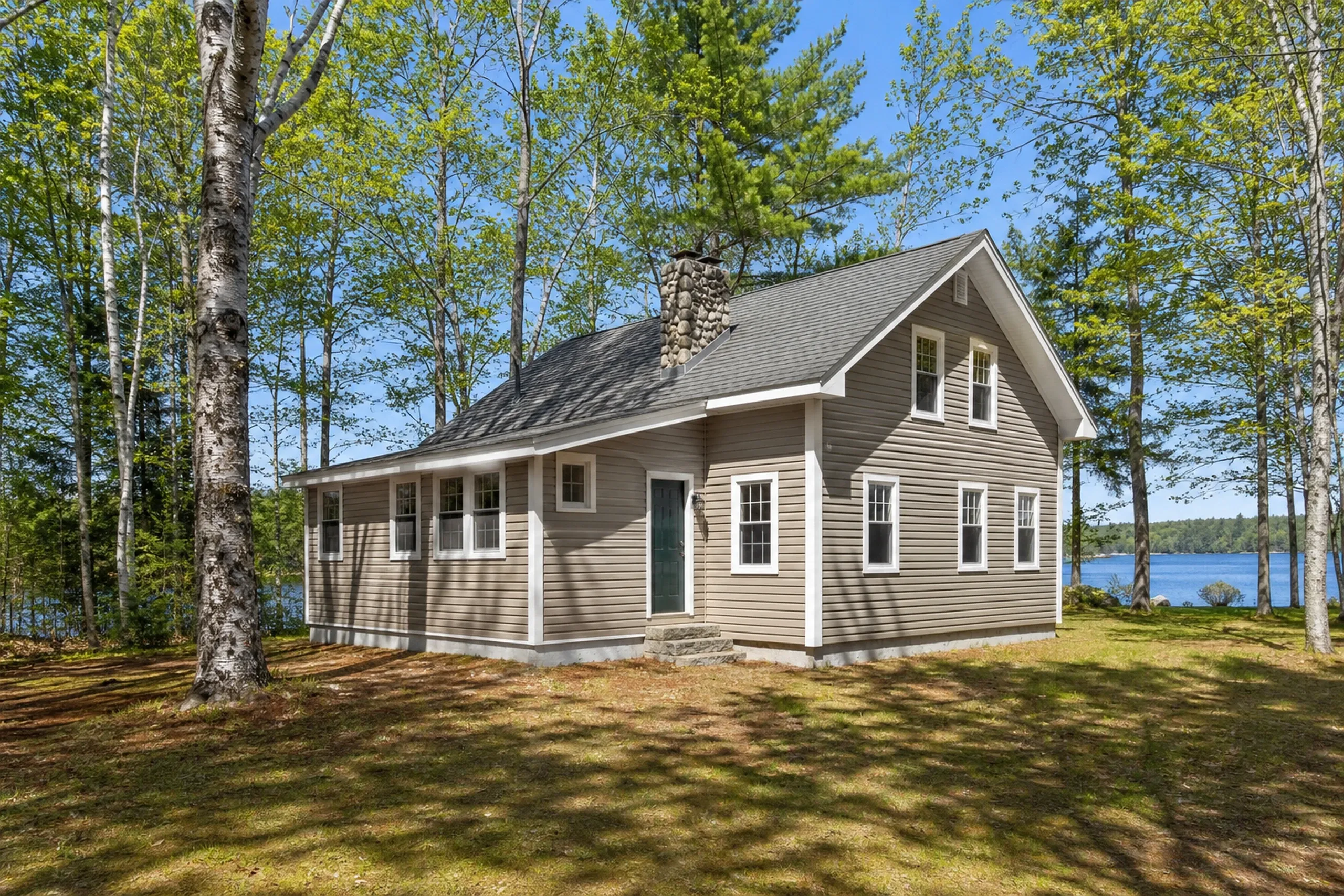 Re-sided Oxford lake camp in early summer: A re-sided year-round camp on a wooded shore of Thompson Lake, finished in soft taupe vinyl lap with white trim and a forest-green door, a fieldstone chimney, white birches and a bright blue slice of lake between the trunks, bright early-summer afternoon light.